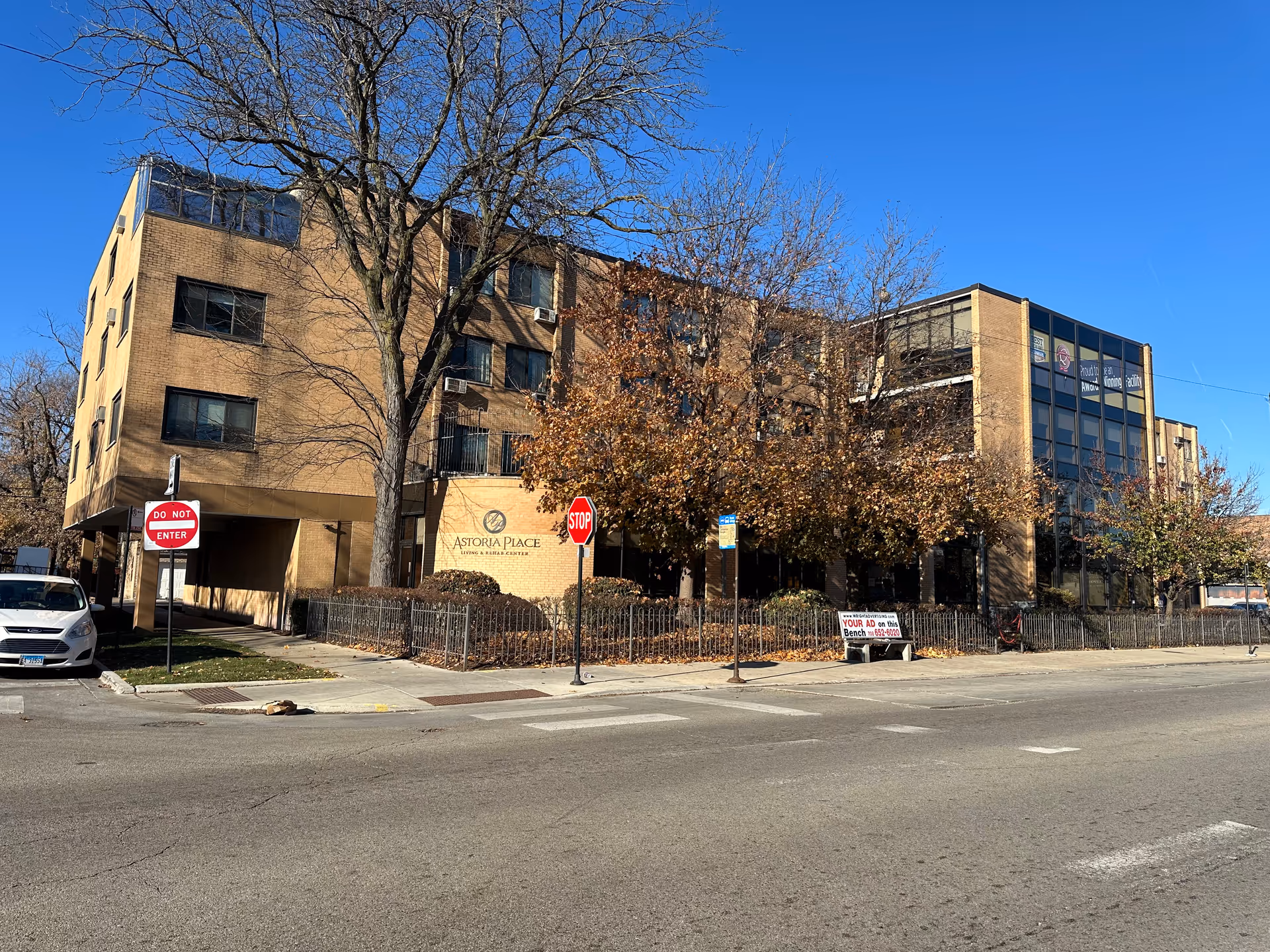 Front exterior of Astoria Place Living & Rehab Center, a multi-story brick building with trees, a fenced entrance and street signs in front.