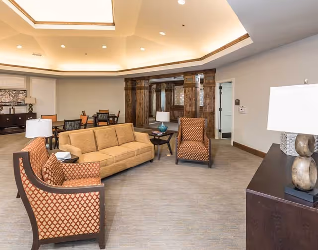 A spacious living room area in a senior living facility with a beige sofa, two patterned armchairs, a side table with a lamp, and a dark wooden cabinet with a decorative lamp. The room has a high ceiling with recessed lighting and wooden architectural details in the background. There are also tables and chairs in the background for additional seating.