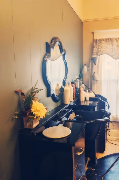 Interior view of a hair salon station with a black sink, a large decorative mirror on the wall, various hair care products on the counter, a small flower arrangement, and a window with a curtain letting in natural light.