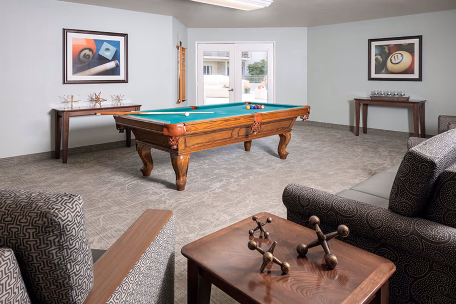 A recreational room with a wooden pool table in the center, two framed pictures of billiard balls on the walls, two wooden tables with decorative items, and patterned armchairs and a sofa around a wooden coffee table.