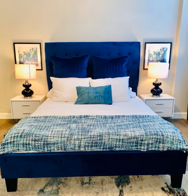 A neatly made bed with a navy upholstered headboard, blue and white pillows, a patterned blue throw, and matching bedside tables with lamps and framed artwork.