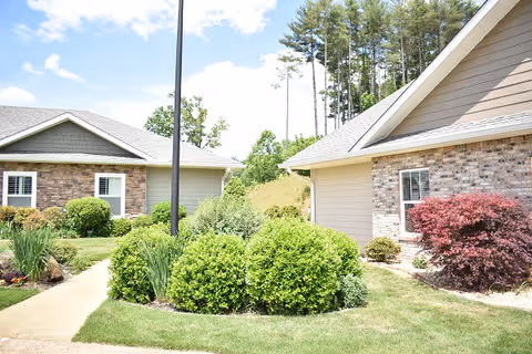 Outdoor view of a senior living facility showing two single-story buildings with beige siding and brick accents, surrounded by well-maintained green bushes, shrubs, and a grassy lawn under a partly cloudy sky.