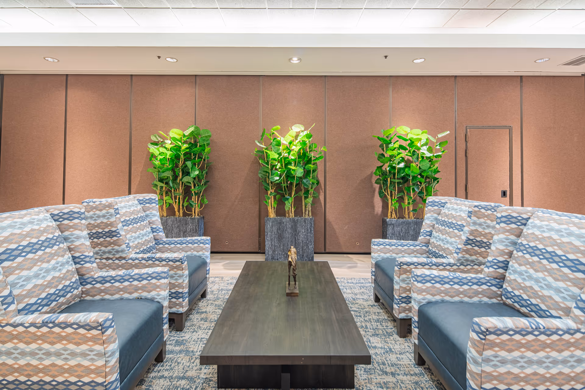 Symmetrical lounge with patterned armchairs facing a central coffee table and tall potted plants against a paneled wall.