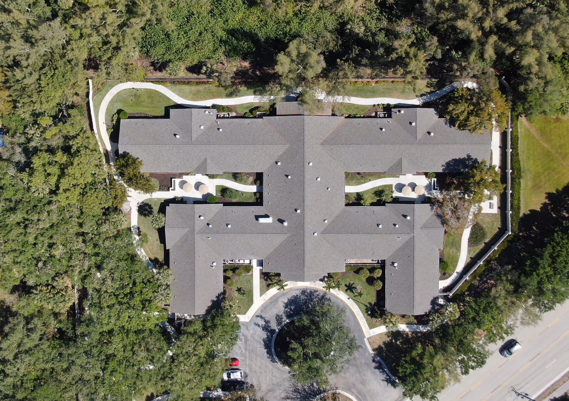 Aerial view of Arden Courts - ProMedica Memory Care Community in Ft. Myers, showing a large building with a complex roof structure surrounded by trees and greenery. There are curved walkways around the building and a circular driveway with parked cars at the entrance.