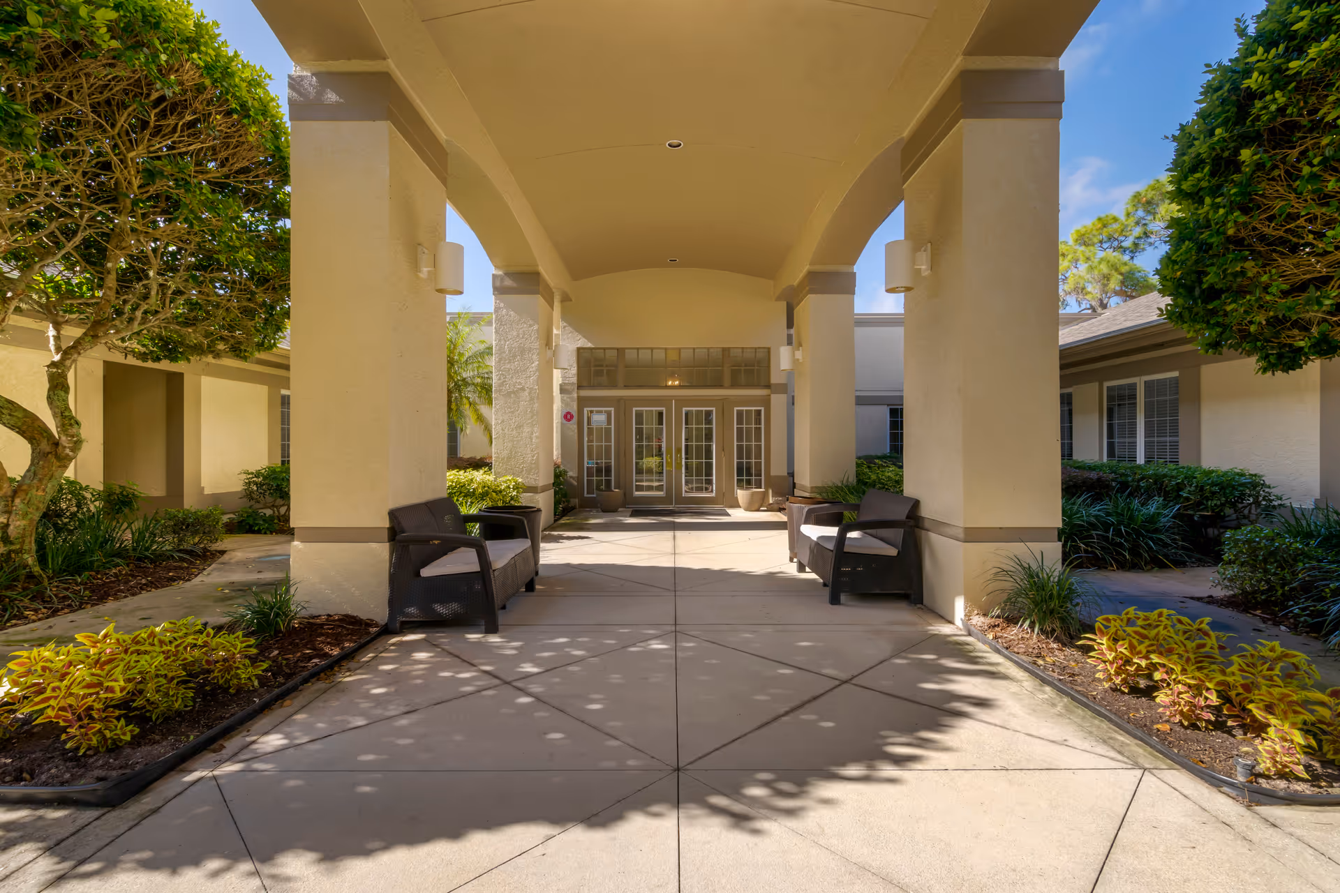 Covered outdoor walkway with beige columns and ceiling leading to glass double doors entrance. There are two black cushioned benches on either side of the walkway and landscaped plants and trees surrounding the area under a clear blue sky.