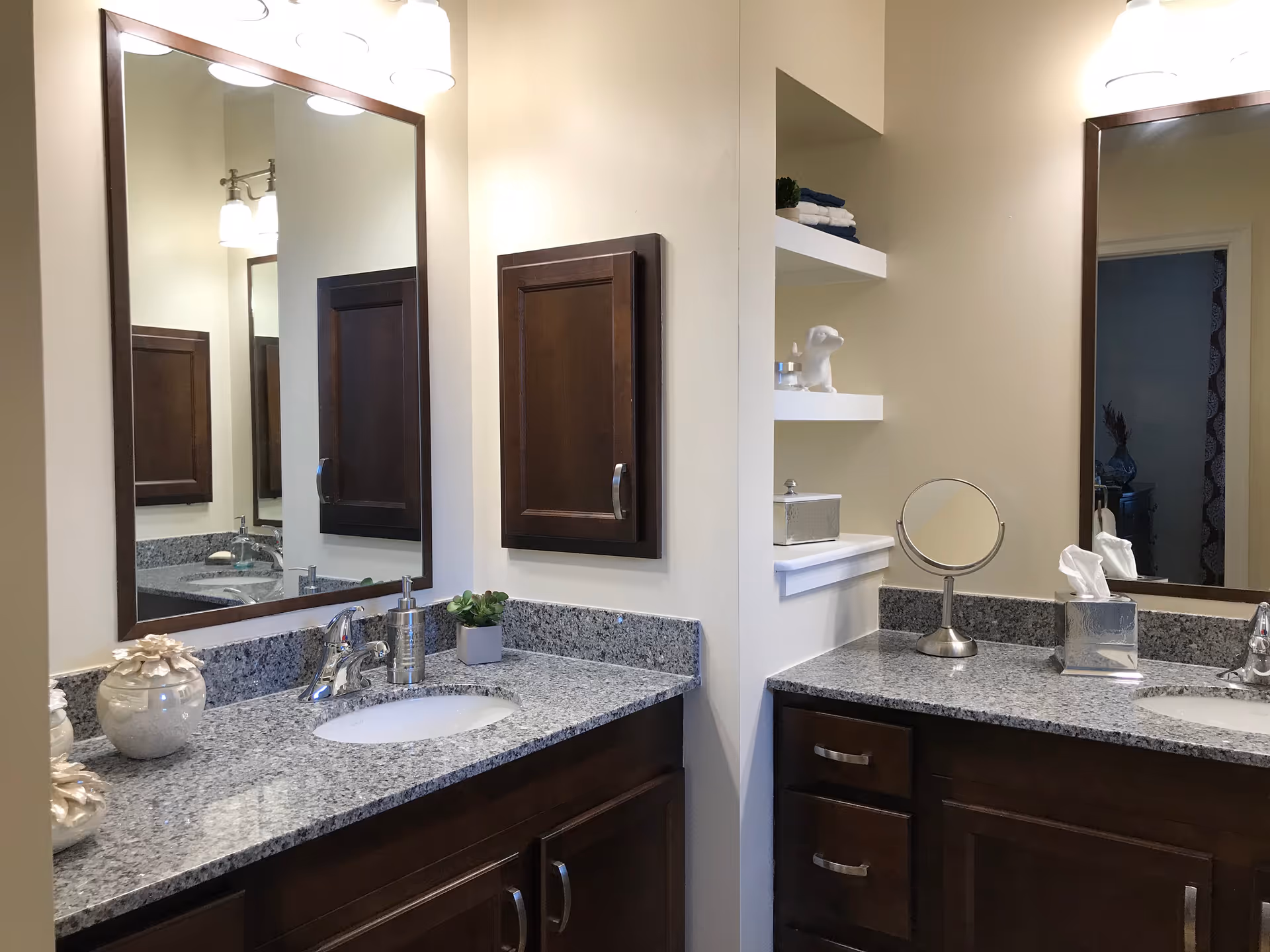 A bathroom with two granite countertops and sinks, each with a large mirror above. The cabinetry is dark wood, and there are decorative items including a small plant, a soap dispenser, a tissue box, and a small round mirror. The walls are light-colored, and there are built-in shelves with towels and decor.