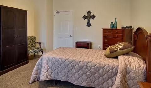 A furnished bedroom with a single bed, wooden dresser and wardrobe, an upholstered chair, and a decorative wall cross.