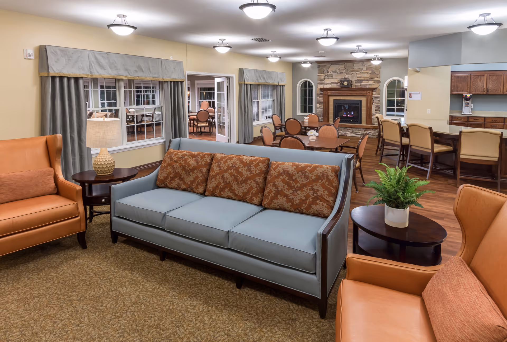 A cozy senior living facility common area with a light blue sofa adorned with patterned cushions, two orange armchairs, a wooden side table with a lamp, and another side table with a potted plant. In the background, there are dining tables with chairs, a stone fireplace, and a kitchen area with bar stools. The room has beige walls, carpeted flooring, and multiple ceiling lights.