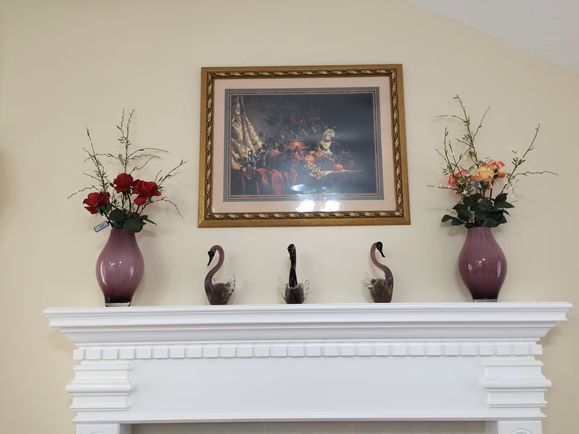 A white mantel topped with two purple vases of flowers, three glass swan figurines, and a framed still-life painting on a beige wall.