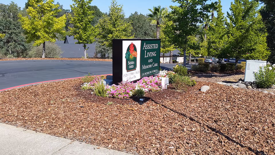 Outdoor landscaped area with a sign for Sierra Oaks Assisted Living and Memory Care surrounded by flowers and mulch, with trees and a parking lot in the background under a clear blue sky.