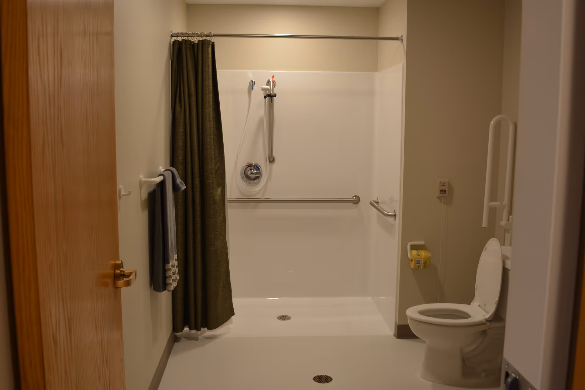 A bathroom with a walk-in shower featuring grab bars and a handheld showerhead. There is a green shower curtain, a towel hanging on a wall hook, a toilet with grab bars on both sides, and a toilet paper holder mounted on the wall. The walls are beige and the floor is light-colored.