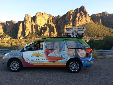 A Doctor's Choice Assisted Living van parked near rugged desert mountains with resident portraits on side.