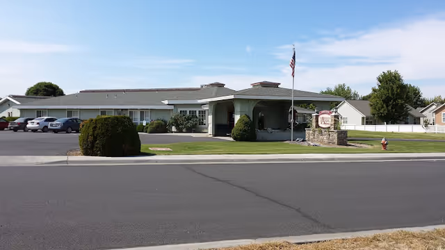 Exterior view of a single-story senior living facility building with a covered entrance, an American flag on a flagpole, a stone sign with the facility name, a parking lot with several cars, and well-maintained landscaping including bushes and grass.