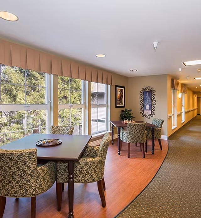 A bright interior common area with large windows letting in natural light. There are two dark wooden tables, each surrounded by four patterned upholstered chairs. The floor is a combination of wood and carpet, and the walls are decorated with framed artwork and a decorative mirror. The space appears cozy and inviting, with a hallway extending to the right.