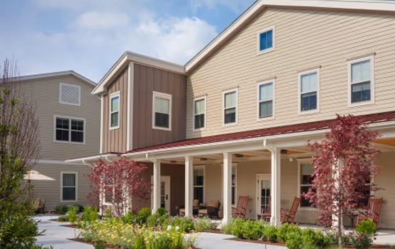 Exterior view of a senior living facility building with beige siding and a covered porch area featuring rocking chairs. There are landscaped gardens with green shrubs and small trees with red leaves in front of the building under a partly cloudy sky.