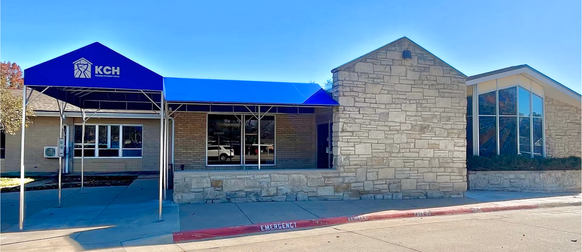 Front entrance of the Kansas Christian Home building with a blue KCH awning, stone facade, and large windows.