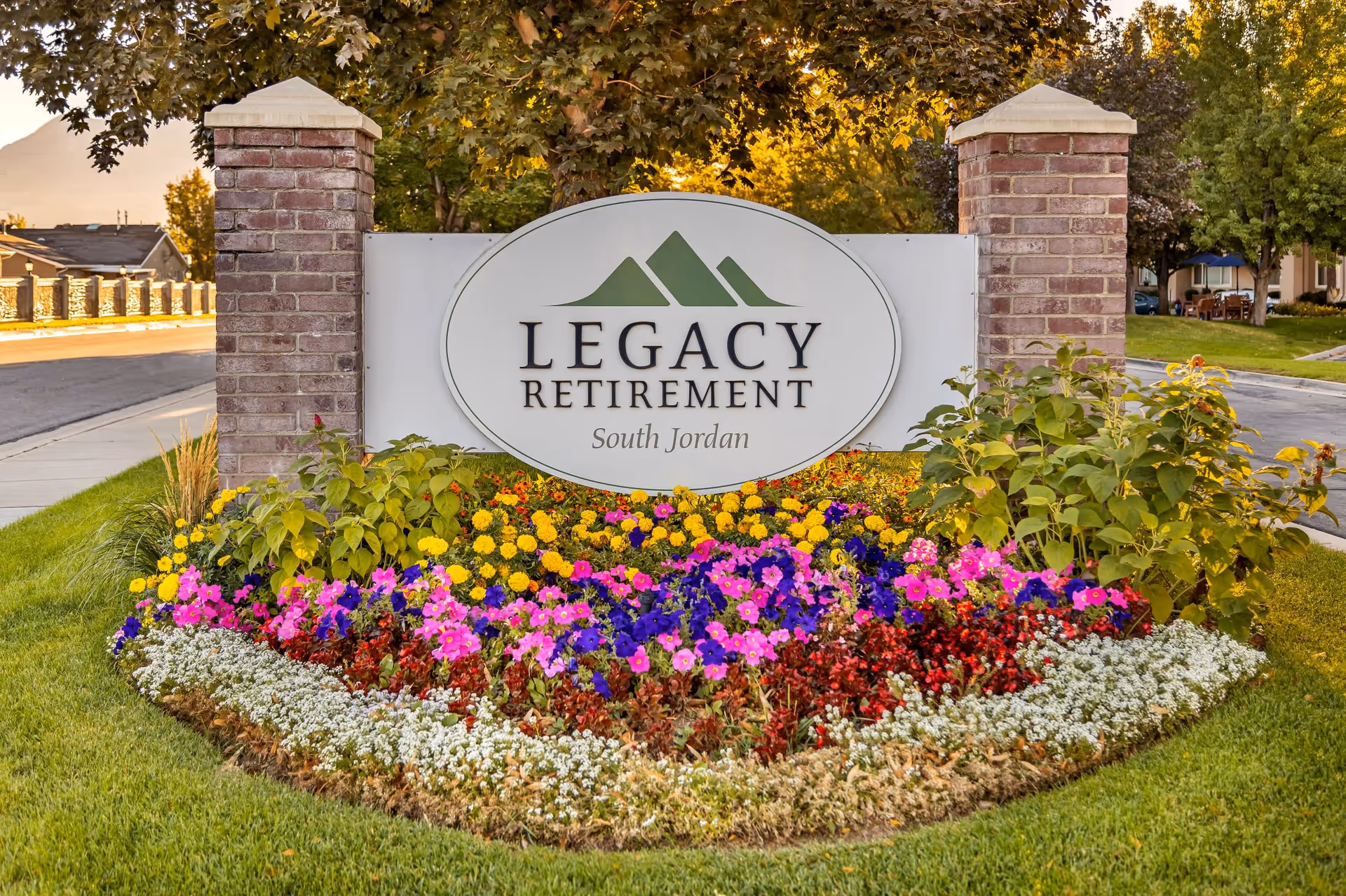 A large sign for Legacy Retirement South Jordan set between two brick pillars, surrounded by a colorful flower bed with various blooming flowers and green plants, with a street and trees in the background.