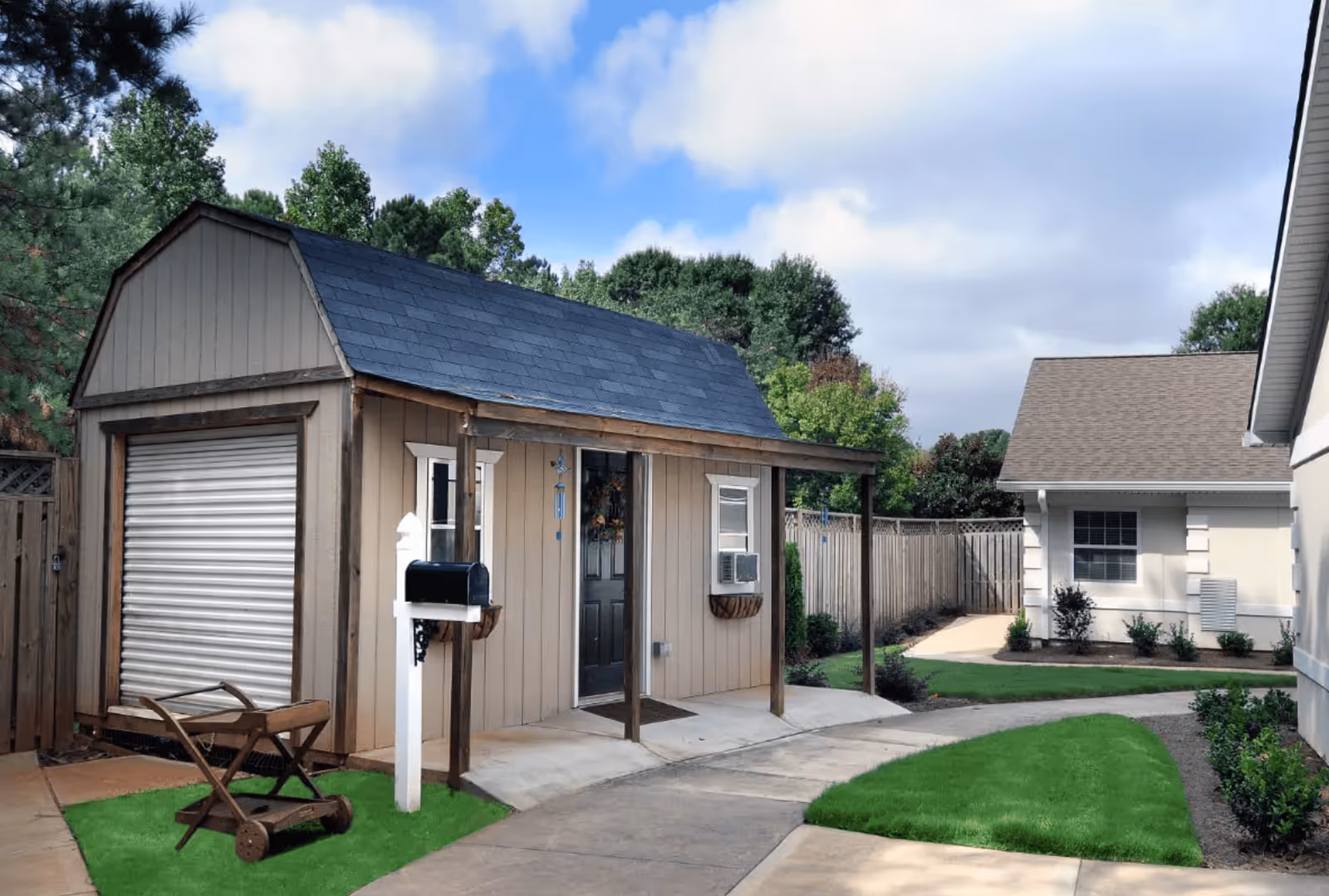 Small beige shed with a black door and a window air conditioning unit, located next to a paved pathway with green grass patches and surrounded by trees and other buildings under a partly cloudy sky.
