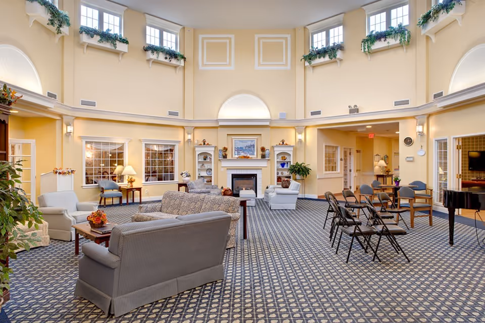 A spacious and well-lit living room area with high ceilings and multiple windows near the ceiling adorned with green plants. The room features several upholstered sofas and armchairs arranged around a central fireplace with built-in shelves on either side. There are also folding chairs set up in rows, a piano on the right side, and various lamps and plants decorating the space. The walls are painted a soft yellow, and the floor is covered with a patterned carpet.