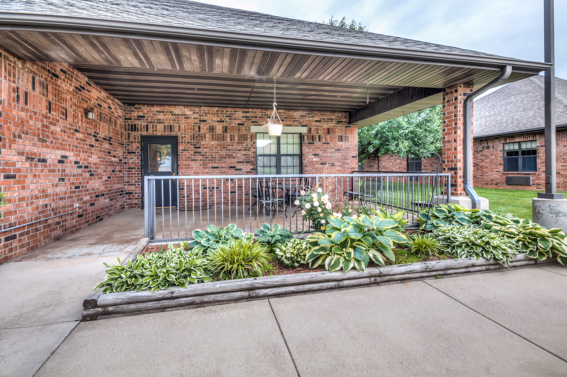 Covered outdoor patio area with brick walls, a black metal railing, and a hanging light fixture. There is a small garden bed with green leafy plants and flowers in front of the patio. In the background, there are more brick buildings and green grass.