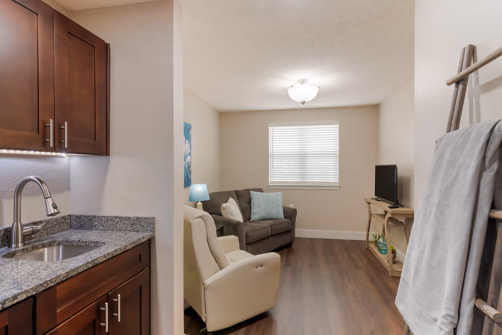 Interior view of a memory care community living space featuring a small kitchenette with granite countertops and dark wood cabinets on the left, a cozy sitting area with a gray loveseat, a white armchair, a small side table with a blue lamp, a TV on a wooden stand, and a window with closed blinds. A wooden ladder with a gray blanket is on the right side of the image.