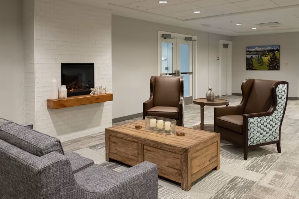 A cozy common lounge with a wall-mounted fireplace, gray sofa, two brown wingback chairs, and a wooden coffee table.