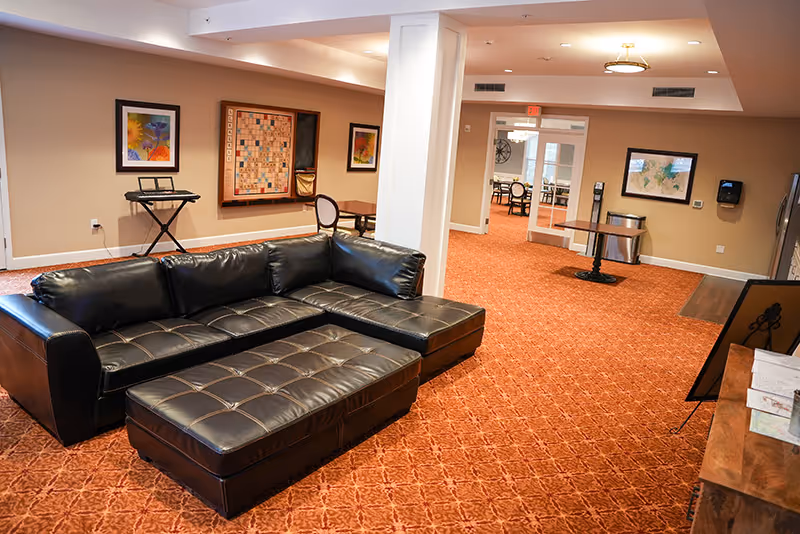 A spacious lounge with a black leather sectional sofa on patterned red carpet, tables and framed artwork in a well-lit common area.