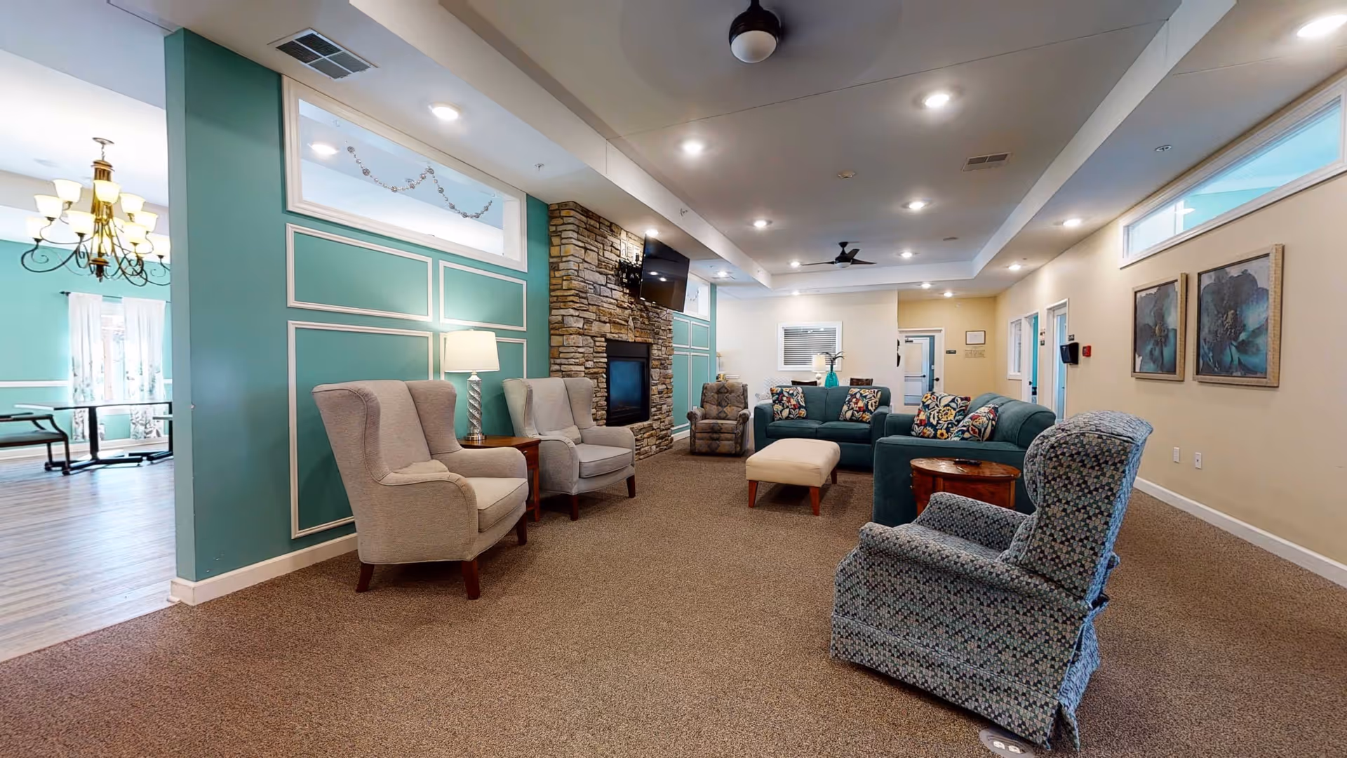 A spacious and well-lit living room area in a retirement living facility featuring comfortable armchairs, a sofa with patterned cushions, a stone fireplace with a mounted TV above it, and soft carpeting. The walls are painted in a combination of teal and beige, with decorative molding and framed artwork. In the background, a dining area with a chandelier and large windows can be seen.