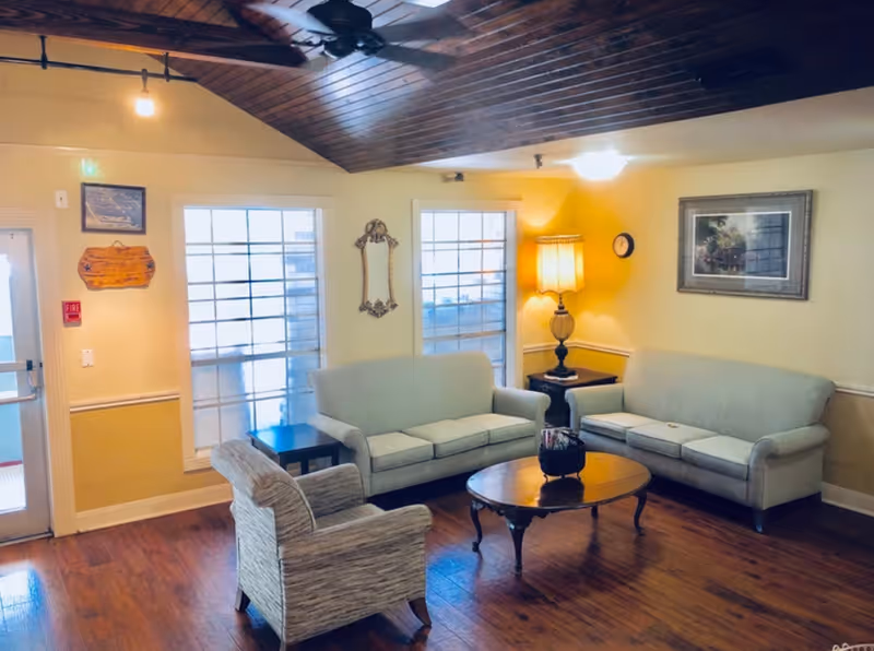 A cozy sitting room with two light-gray sofas, an armchair, a wooden coffee table, a lamp, and a wood-paneled ceiling with a ceiling fan.