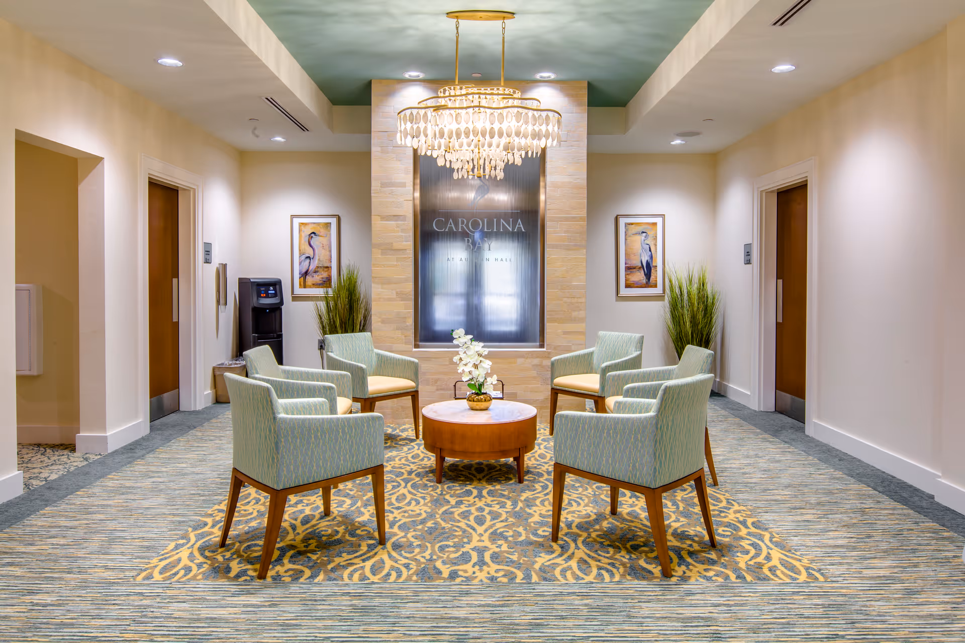 A seating area in a senior living facility with six light green upholstered chairs arranged around a round wooden table with a small floral arrangement. The area has a patterned carpet and neutral-colored walls with two framed bird paintings and tall green plants on either side of a decorative water feature with the text 'Carolina Bay at Autumn Hall'. A chandelier with hanging shells is suspended from the ceiling above the seating area.