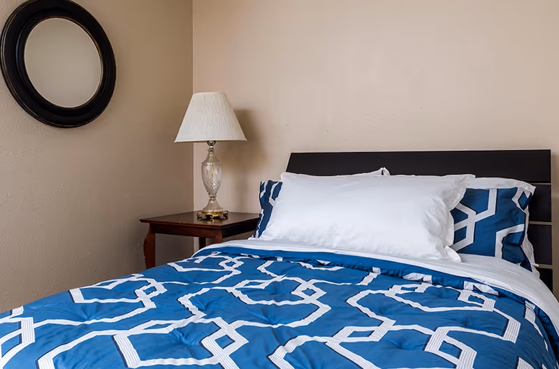 Neatly made bed with blue geometric bedding, white pillows, a dark headboard, bedside table with lamp, and a round wall mirror.