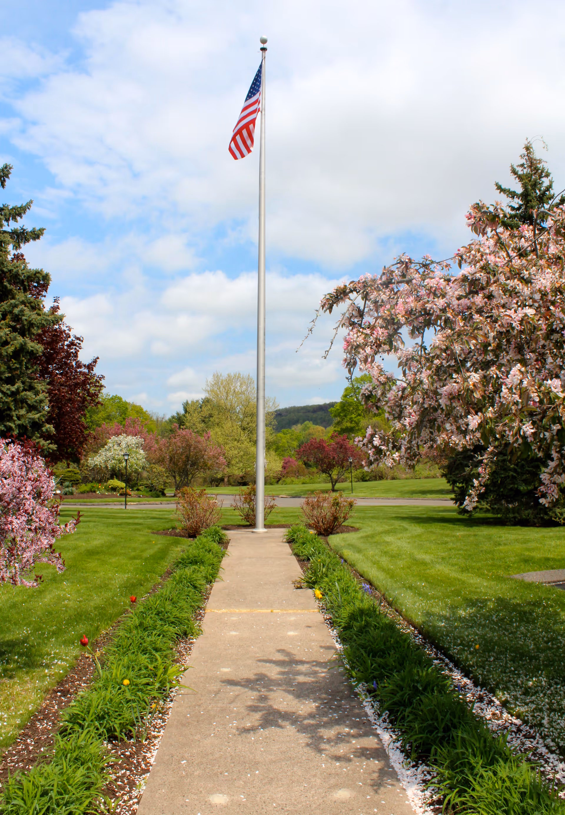 A concrete pathway lined with green plants and flowers leads to a tall flagpole flying the American flag. The surrounding area features well-maintained green lawns, blooming trees with pink and white flowers, and a partly cloudy sky.