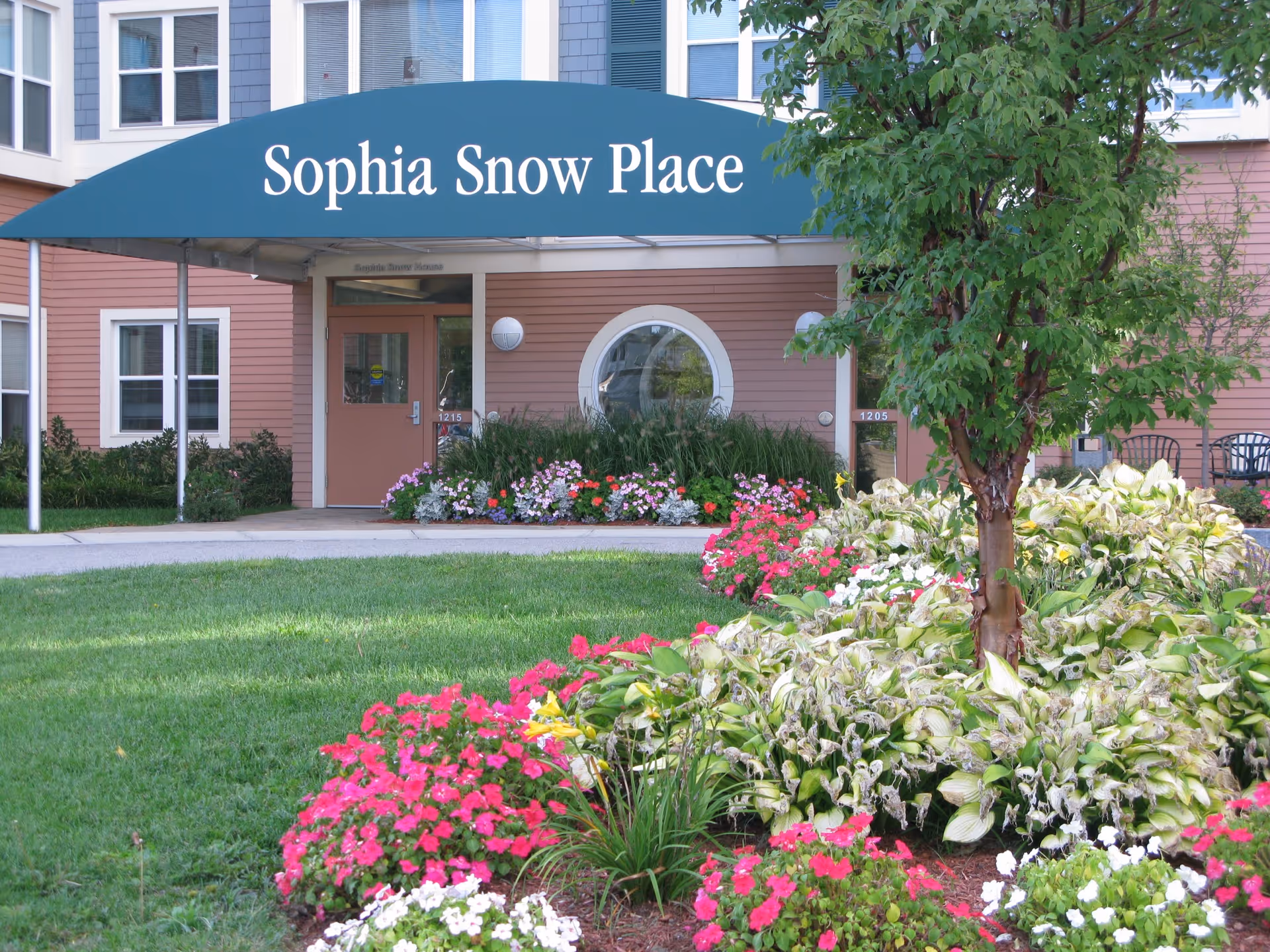 Front entrance of Sophia Snow Place with a green awning, pink building facade, and colorful flower beds in the foreground.