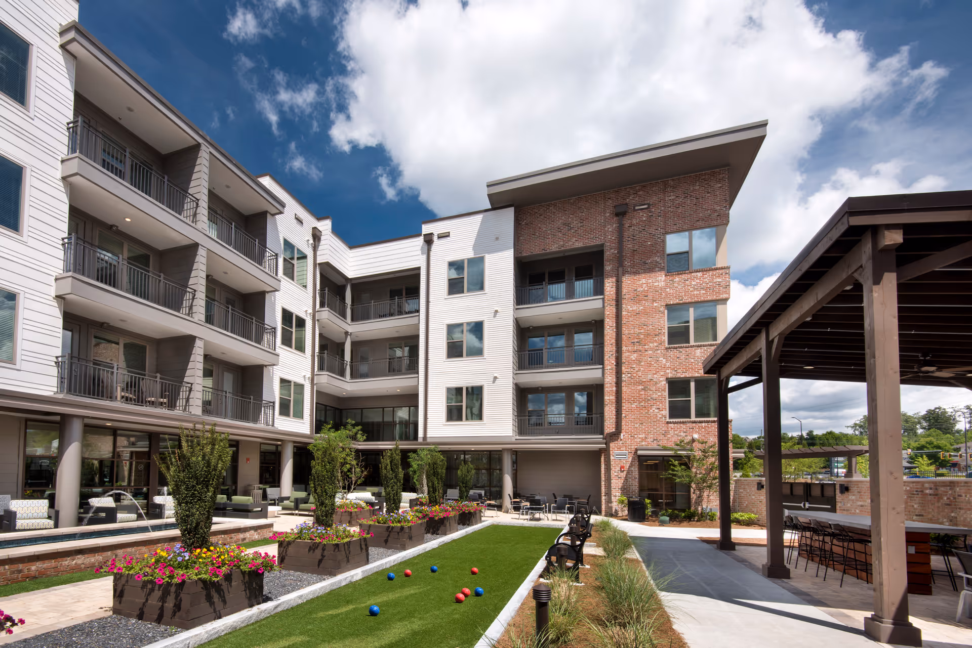 Outdoor courtyard area of a senior living facility with a bocce ball court, flower planters, benches, and a covered seating area. The building has multiple floors with balconies and a mix of white siding and red brick exterior under a partly cloudy sky.