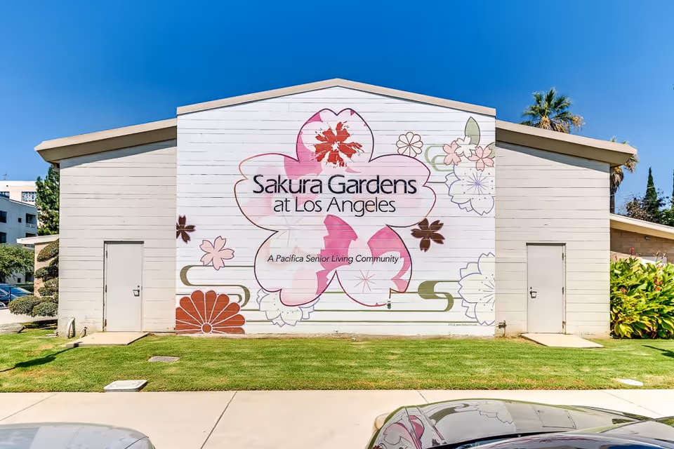 Exterior view of a building with a large mural featuring pink and white flowers and the text 'Sakura Gardens at Los Angeles, A Pacifica Senior Living Community'. The building has two closed doors on either side of the mural, a well-maintained lawn in front, and a clear blue sky above.