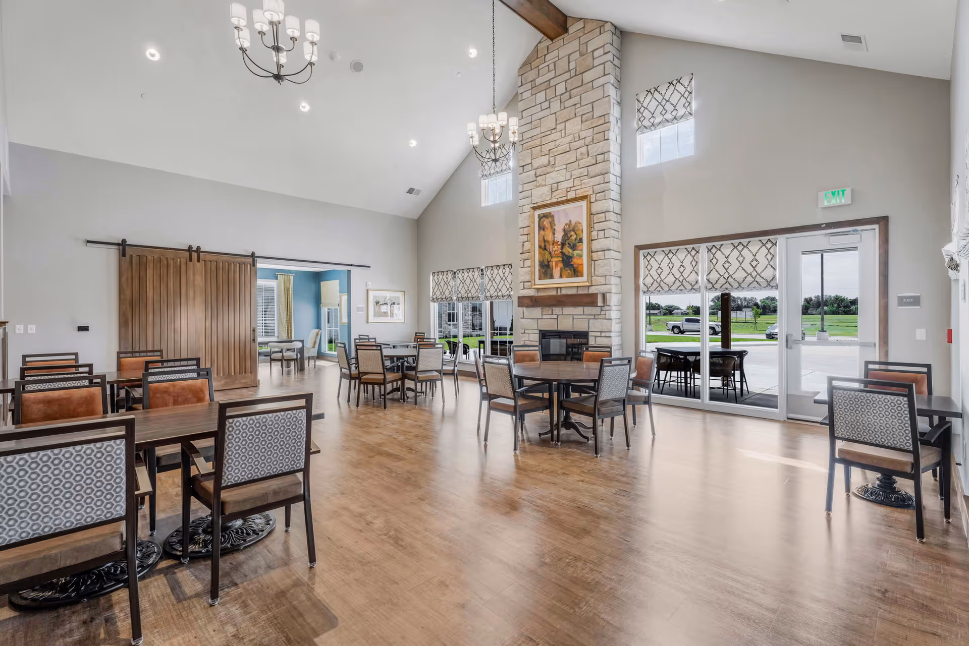 Spacious senior living dining area with multiple tables and chairs, a tall stone fireplace with a painting above it, large windows with patterned valances, sliding barn doors leading to another room, and glass doors opening to an outdoor patio with a view of green grass and trees.