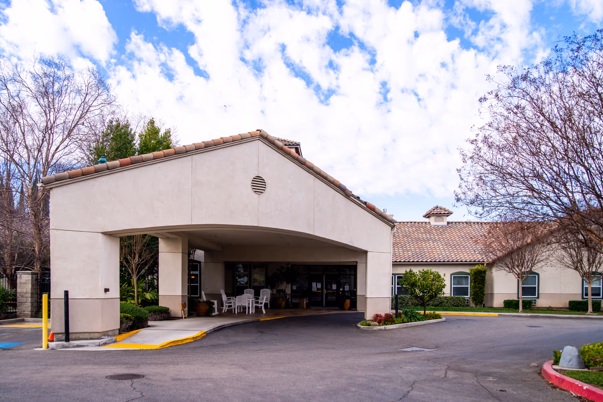 Front exterior view of Astoria Senior Living Tracy building with a covered entrance, outdoor seating with white chairs and tables, surrounded by trees and shrubs under a partly cloudy sky.