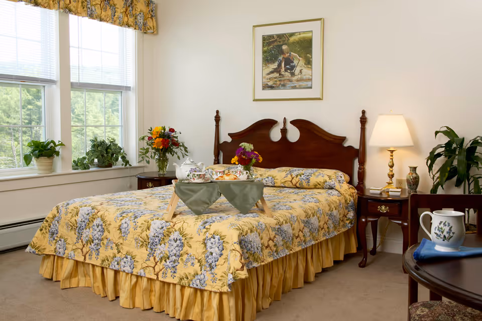 Sunlit bedroom with a wooden headboard, yellow floral bedspread and a breakfast tray, flanked by bedside tables, a lamp and houseplants.