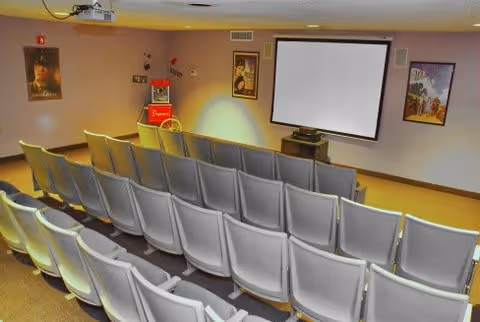 Small movie theater room with rows of plastic chairs facing a projection screen, a popcorn machine, and movie posters on the walls.