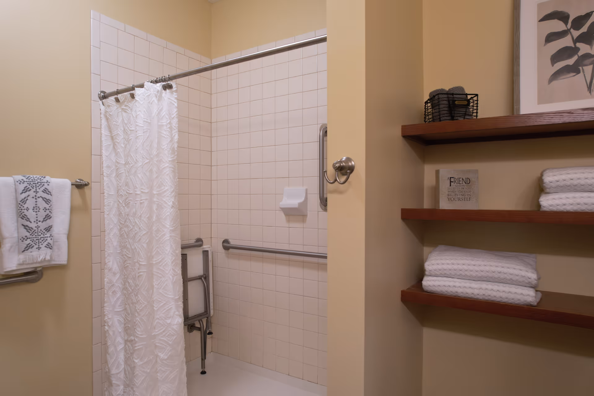 A bathroom shower area with beige tiled walls and a white textured shower curtain. There are metal grab bars installed on the walls for support, a foldable shower seat, and a soap holder. To the right, there are wooden shelves holding neatly folded white towels, a small basket with rolled towels, and a framed picture with a plant design. A towel with a decorative pattern hangs on a metal towel rack on the left wall.