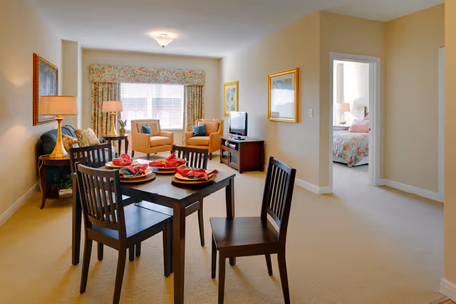 A bright and cozy living and dining area in a senior living facility. The room features a dark wooden dining table set with four chairs and place settings with red napkins. In the background, there are two beige armchairs with blue cushions near a large window with floral curtains. A small TV sits on a wooden stand against the wall. To the right, an open doorway leads to a bedroom with a floral bedspread and a bedside lamp.