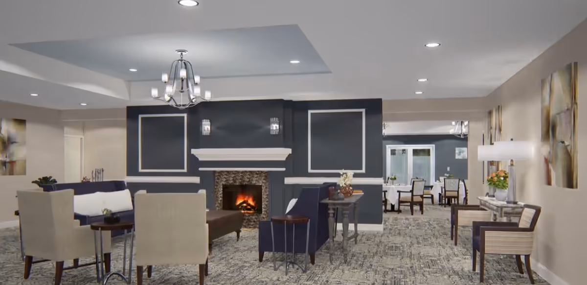 A spacious and well-lit common area with a modern fireplace in the center. The room features comfortable seating including beige and navy chairs around small tables, a chandelier hanging from the ceiling, and decorative wall art. In the background, there is a dining area with tables and chairs set up for meals.