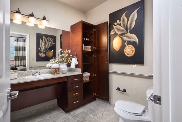 A clean and well-lit bathroom with a granite countertop sink, dark wood cabinets with open doors showing towels and toiletries, a toilet with a grab bar beside it, a large mirror above the sink, and a decorative painting of lemons on the wall.