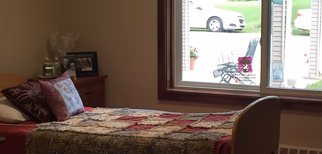 A bedroom with a single bed covered by a patchwork quilt, a wooden nightstand with framed photos and a candle, and a large window showing a view of parked cars and outdoor chairs.