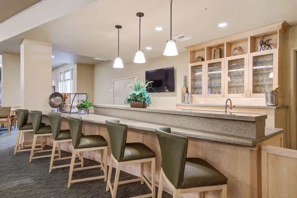Interior view of a senior living facility's bar area with a long counter, six green cushioned bar stools, pendant lights hanging from the ceiling, a wall-mounted TV, and glass cabinets displaying glassware. There are decorative plants on the counter and a carpeted floor.