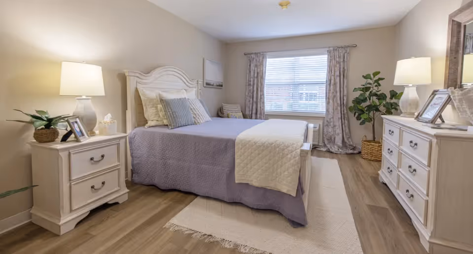 A cozy bedroom with a neatly made bed featuring a lavender quilt and white throw blanket. The room includes two white nightstands with lamps, framed photos, and a plant. A large window with floral curtains lets in natural light, and a white dresser with a mirror and additional decor is on the right side. The floor is wood with a light-colored rug partially under the bed.