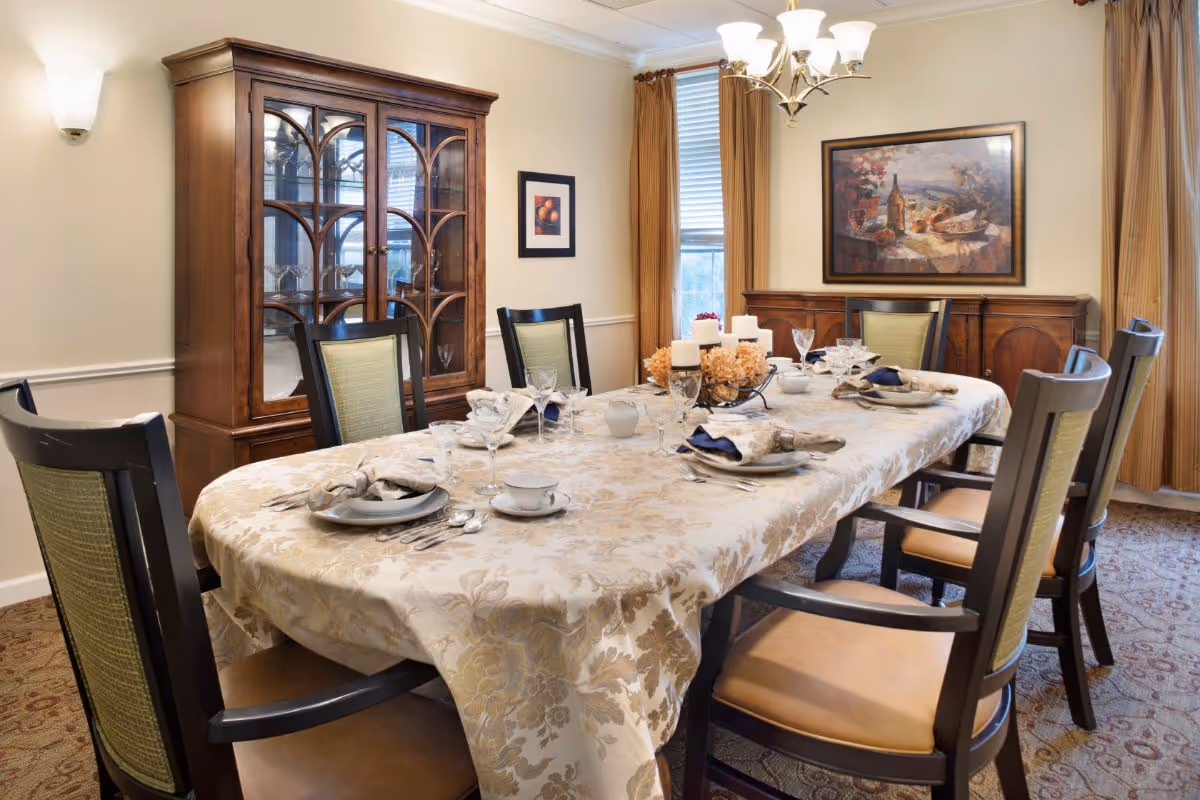 A formal dining room with a long table covered by a floral-patterned tablecloth set for six people with plates, napkins, glasses, and silverware. The room features wooden chairs with green upholstery, a wooden china cabinet with glass doors, a sideboard, a window with gold curtains, a chandelier, and framed artwork on the walls.