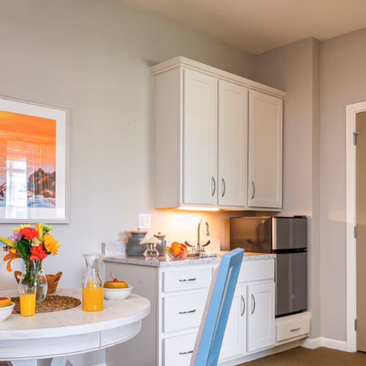 A bright kitchen corner featuring white cabinets, a small stainless steel refrigerator, and a countertop with a sink. A round white table is set with a vase of colorful flowers, a glass pitcher of orange juice, two bowls of fruit, and a glass of orange juice. A blue chair is partially visible in front of the table. A framed artwork with an orange and blue landscape hangs on the wall.