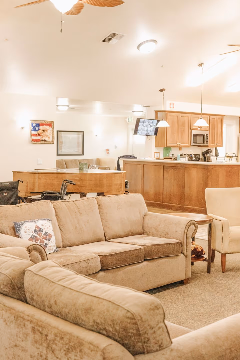A cozy senior living facility common area with beige sofas, a beige armchair, a wooden coffee table, and a grand piano. In the background, there is a kitchen area with wooden cabinets, a microwave, and pendant lights. The walls are decorated with framed pictures and an American flag with an eagle. A wheelchair is visible near the piano.