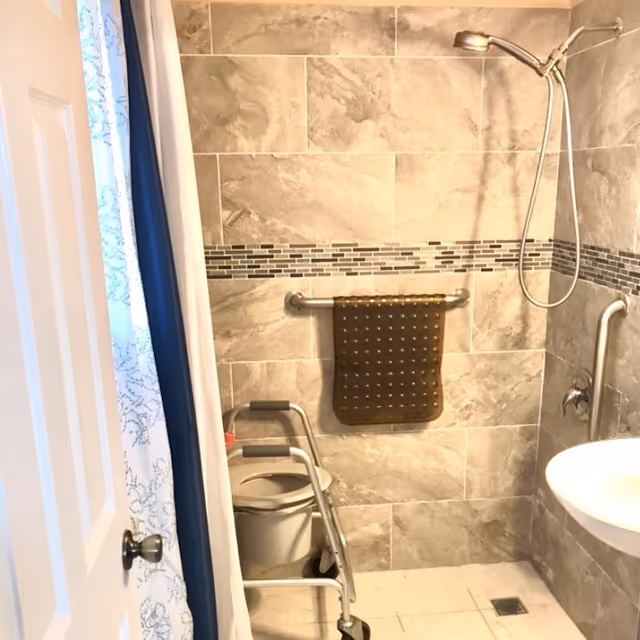 Accessible tiled shower with grab bars, a handheld showerhead, wall-mounted sink, and a wheeled commode/shower chair.